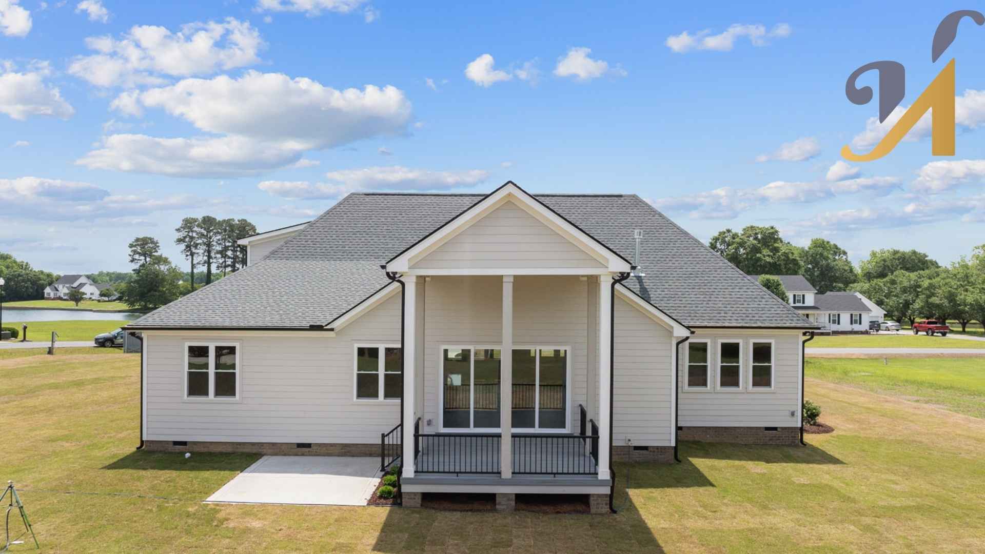 Rear view of a modern, single-story white house with a gabled roof, small back porch with black railings, and freshly landscaped yard under a blue sky with scattered clouds. A lake and a neighboring home are visible in the background. No identifiable people or private details are visible.